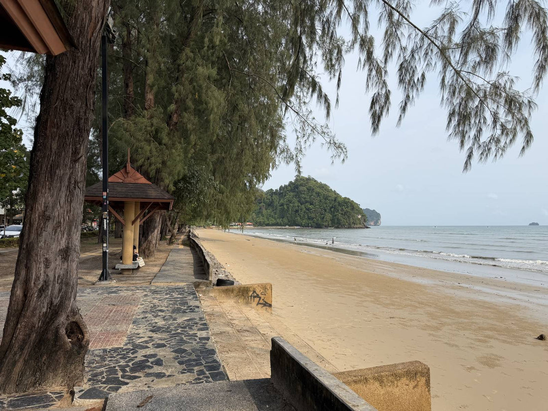 Tree-shaded beachfront walkway along the sand in Ao Nang, Krabi, with an island offshore